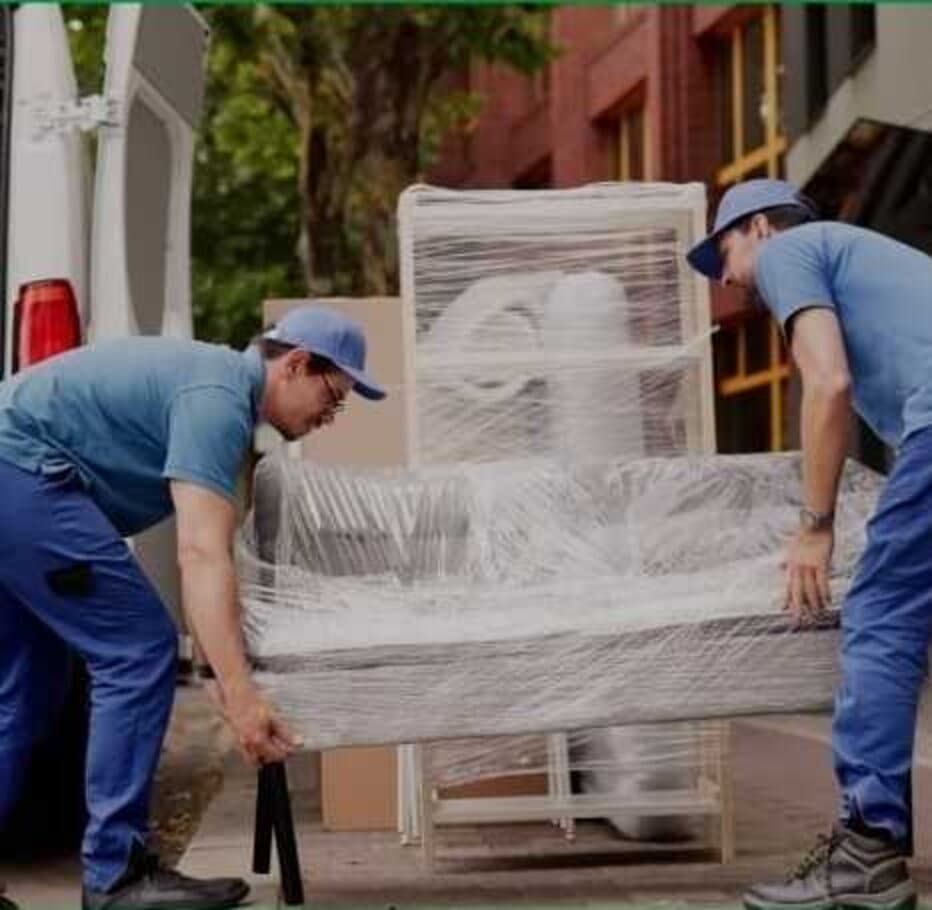 Zwei Männer in blauer Arbeitskleidung verladen ein in Folie eingewickeltes Möbelstück in einen Transporter